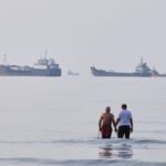 Two men wade in the waters of the Strait of Hormuz with vessels anchored in the background, off Bandar Abbas, Iran, Sunday, July 12, 2026. (Razieh Poudat/ISNA via AP)